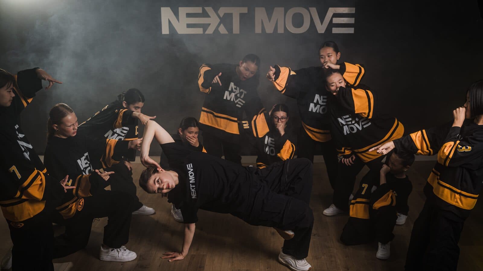 A dancer holds a low breaking pose in the haze while young dancers in Next Move jerseys stand around her.