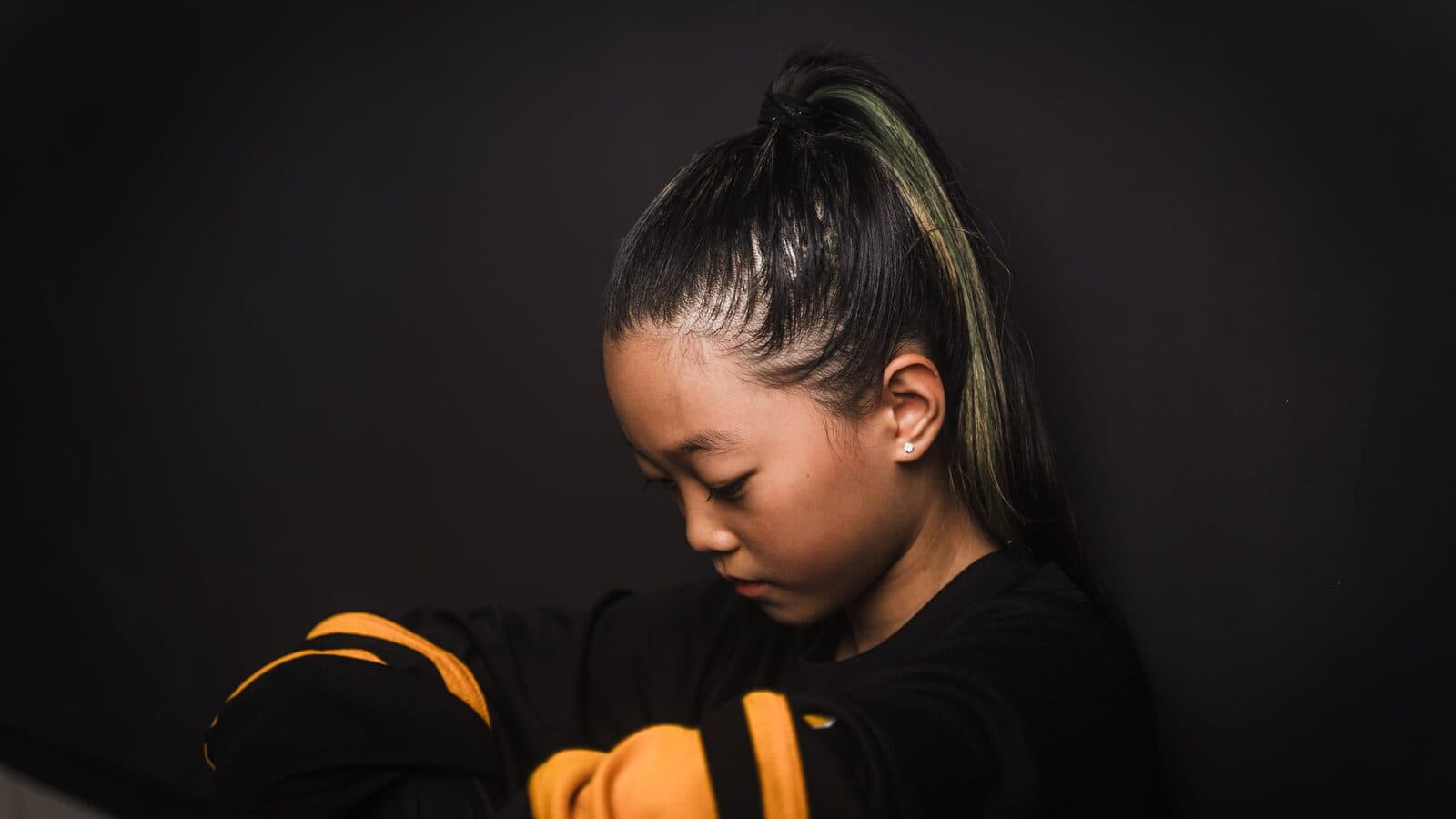 A young dancer looks down in quiet focus against a dark wall.