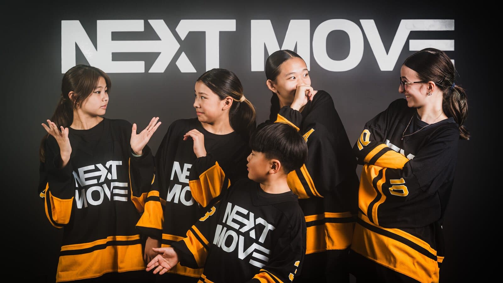Five young dancers in Next Move jerseys shrug, laugh and point in front of the logo wall.