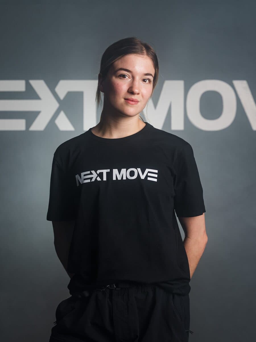 In a Next Move tee, hands behind her back, in front of the studio's logo wall.