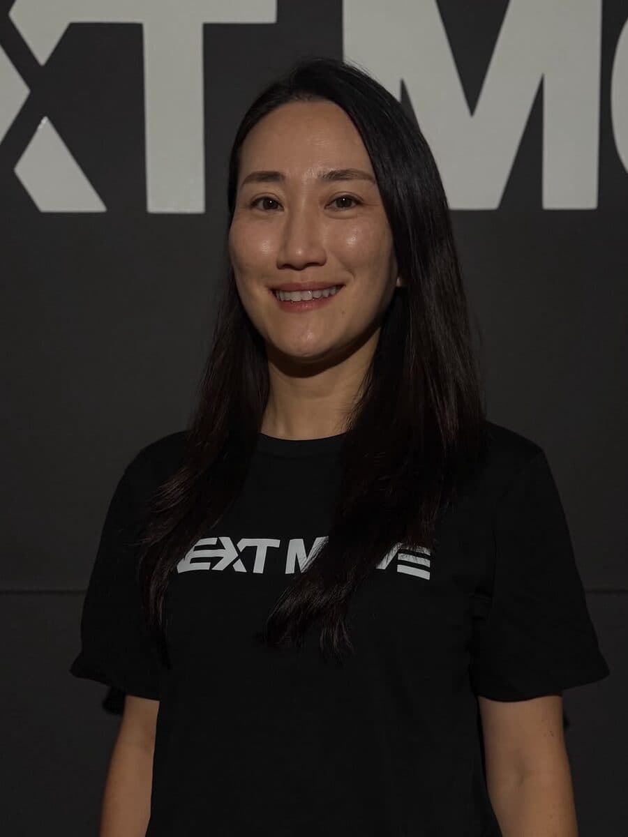 Smiles in a black Next Move tee, arms at her sides, in front of the studio's logo wall.