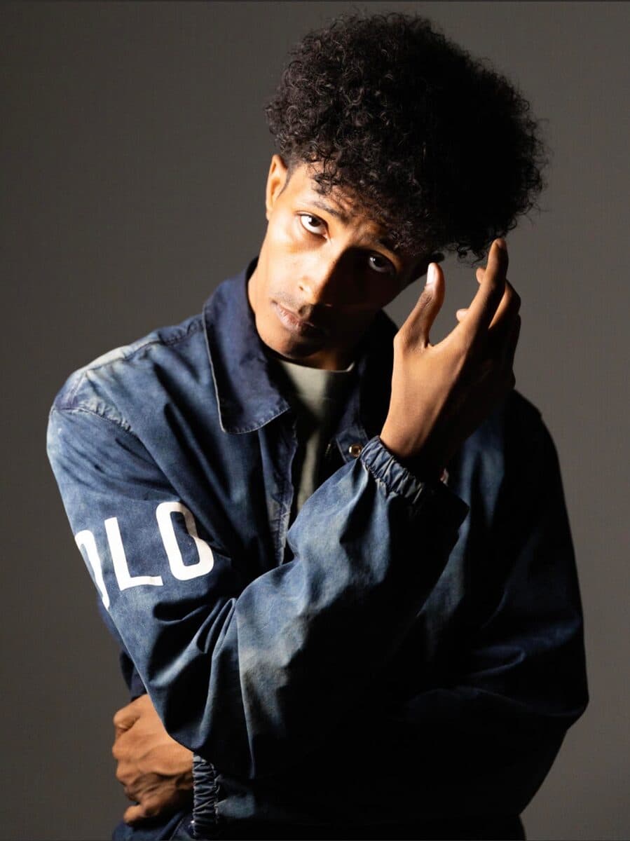 In a denim jacket, one hand at the head, on a dark grey ground.