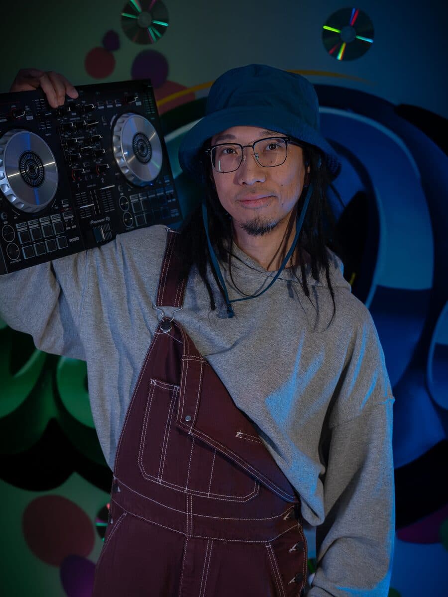 In a bucket hat, glasses and red overalls, a DJ controller on one shoulder, in front of a painted wall.