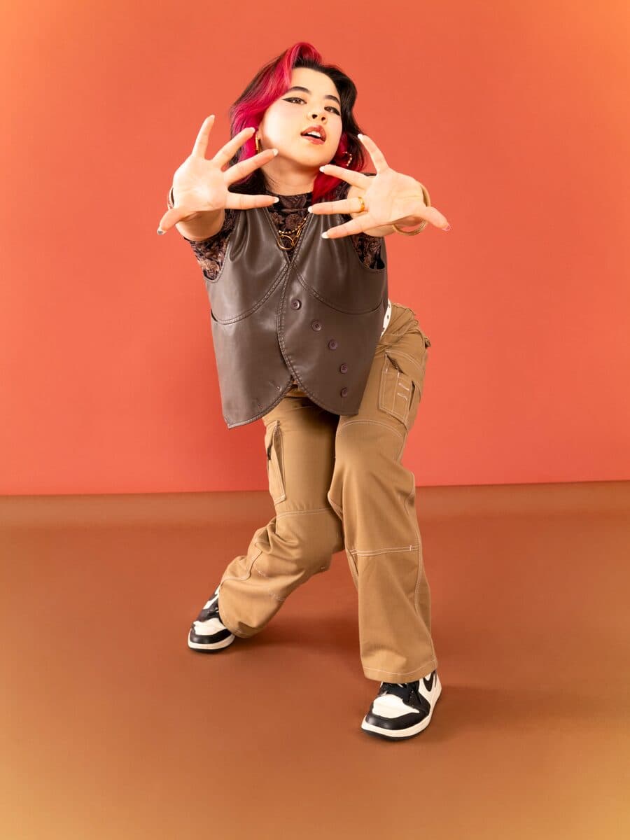 In a leather vest and tan cargo pants, low in a dance stance, hands open to the camera.
