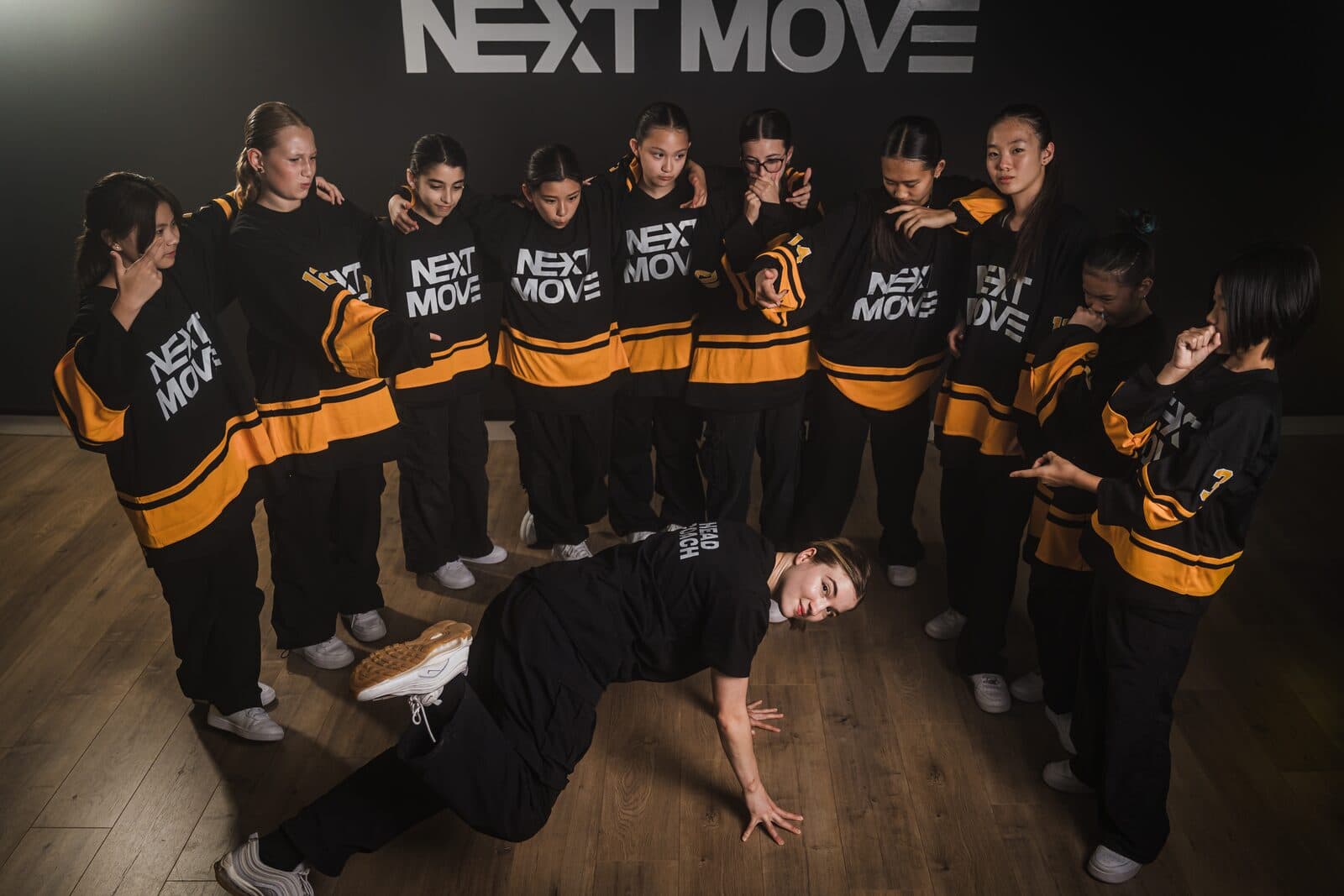 A dancer holds a low one-handed breaking pose on the floor while young dancers in Next Move jerseys stand in a half circle behind her.