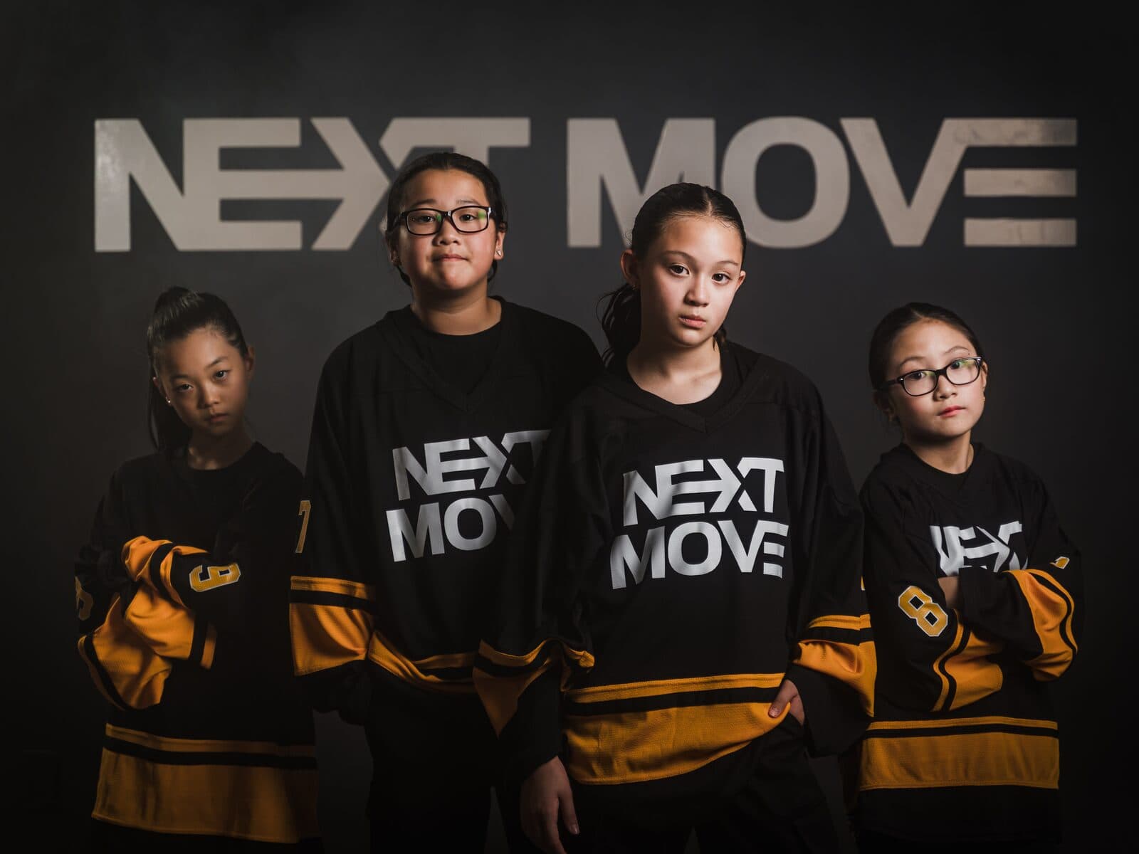 Four young dancers in Next Move jerseys stand close together in front of the studio's logo wall.