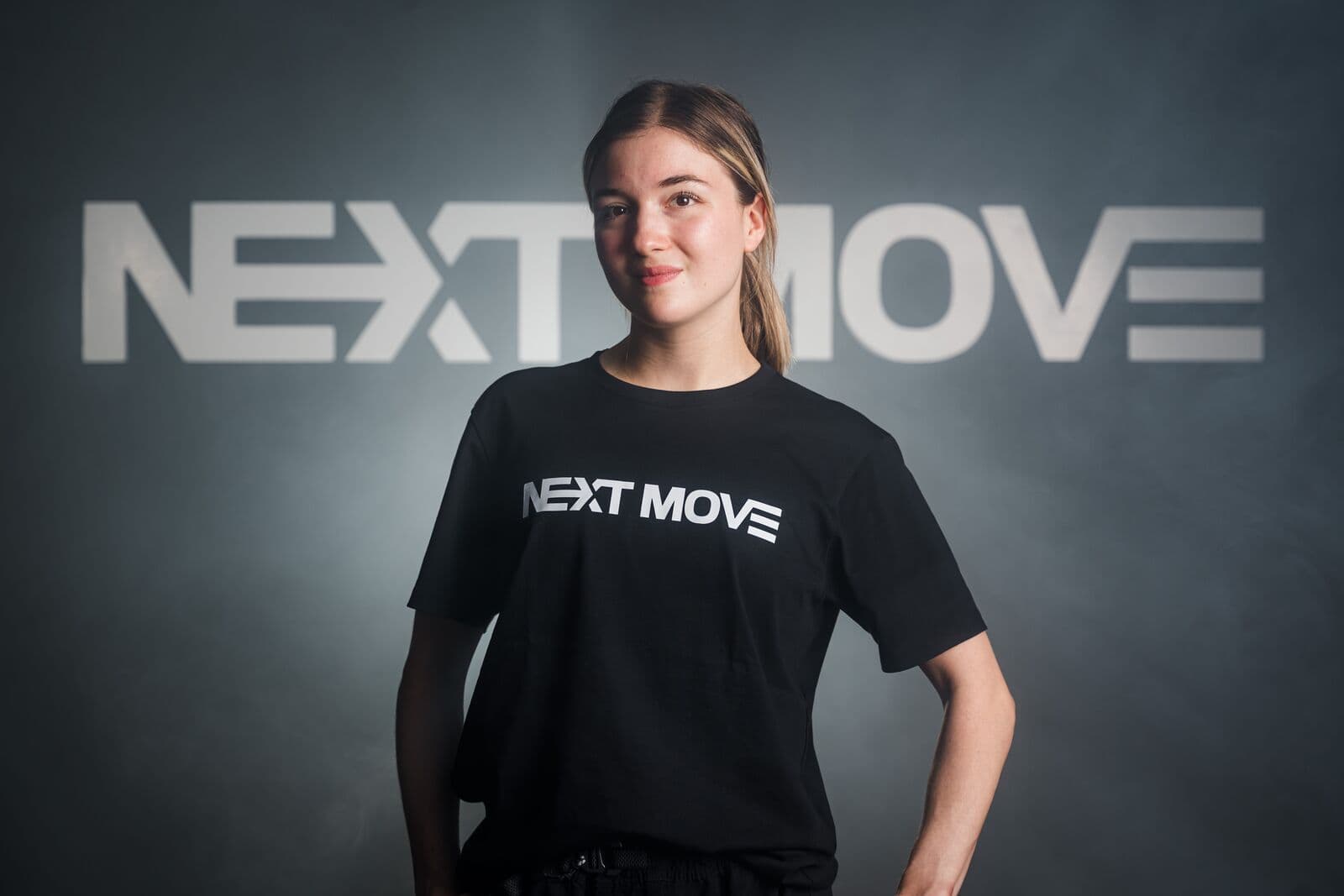 A mentor in a Next Move tee stands in front of the studio's logo wall, one hand on her hip, smiling.