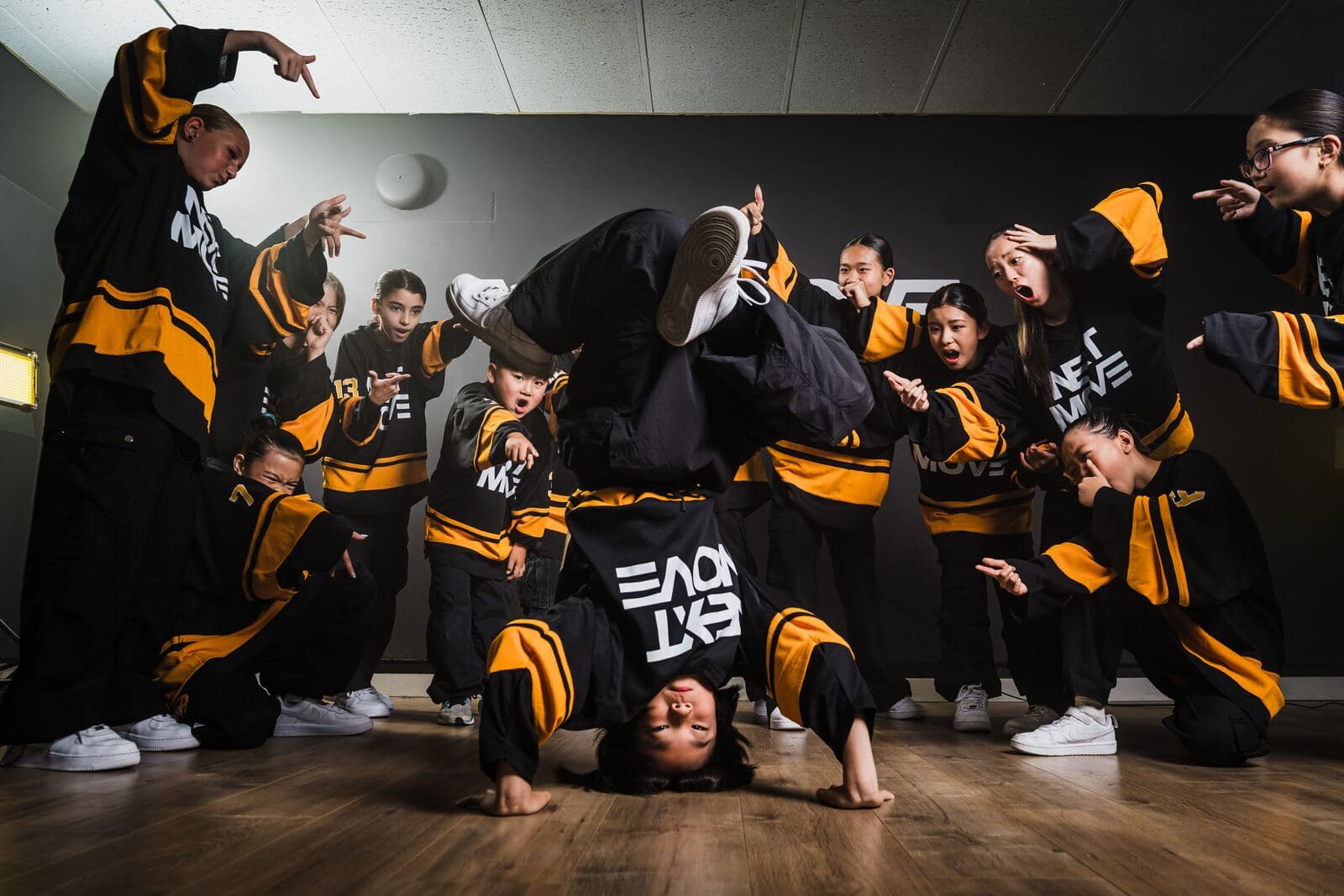 A young breaker holds a headstand while a crew in Next Move jerseys points and reacts around them.
