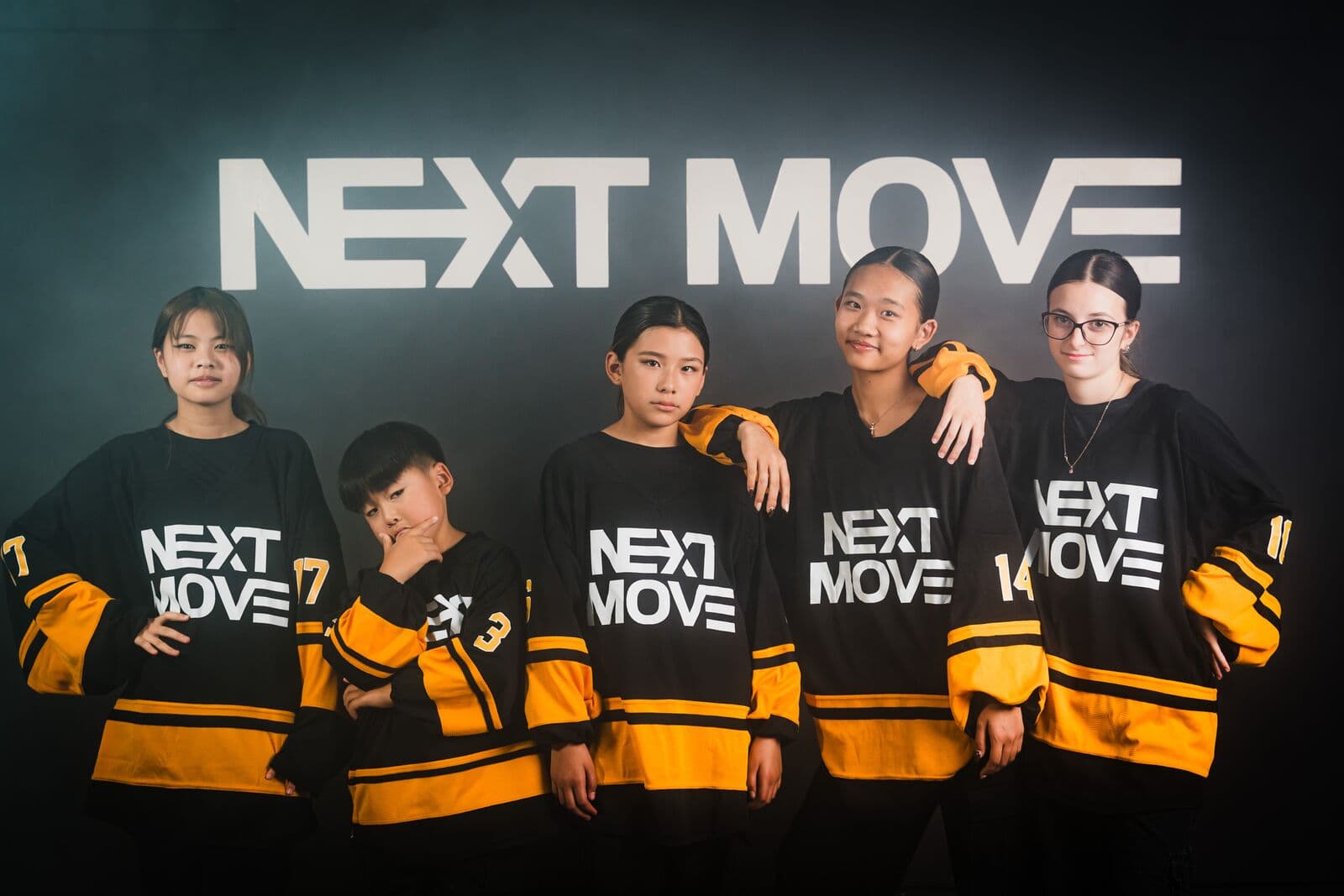 Five young dancers in Next Move jerseys stand in a row in front of the studio's logo wall, two resting an arm on the shoulder of the dancer beside them.