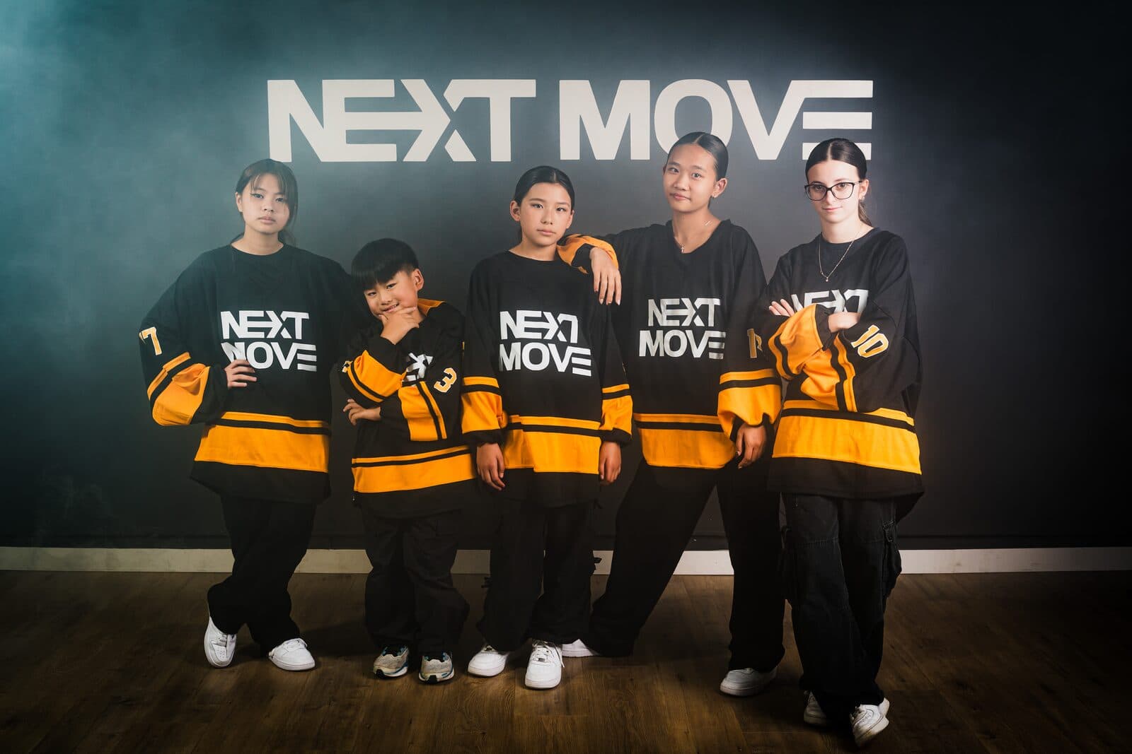 Five young dancers in Next Move jerseys stand in a line in front of the studio's logo wall.