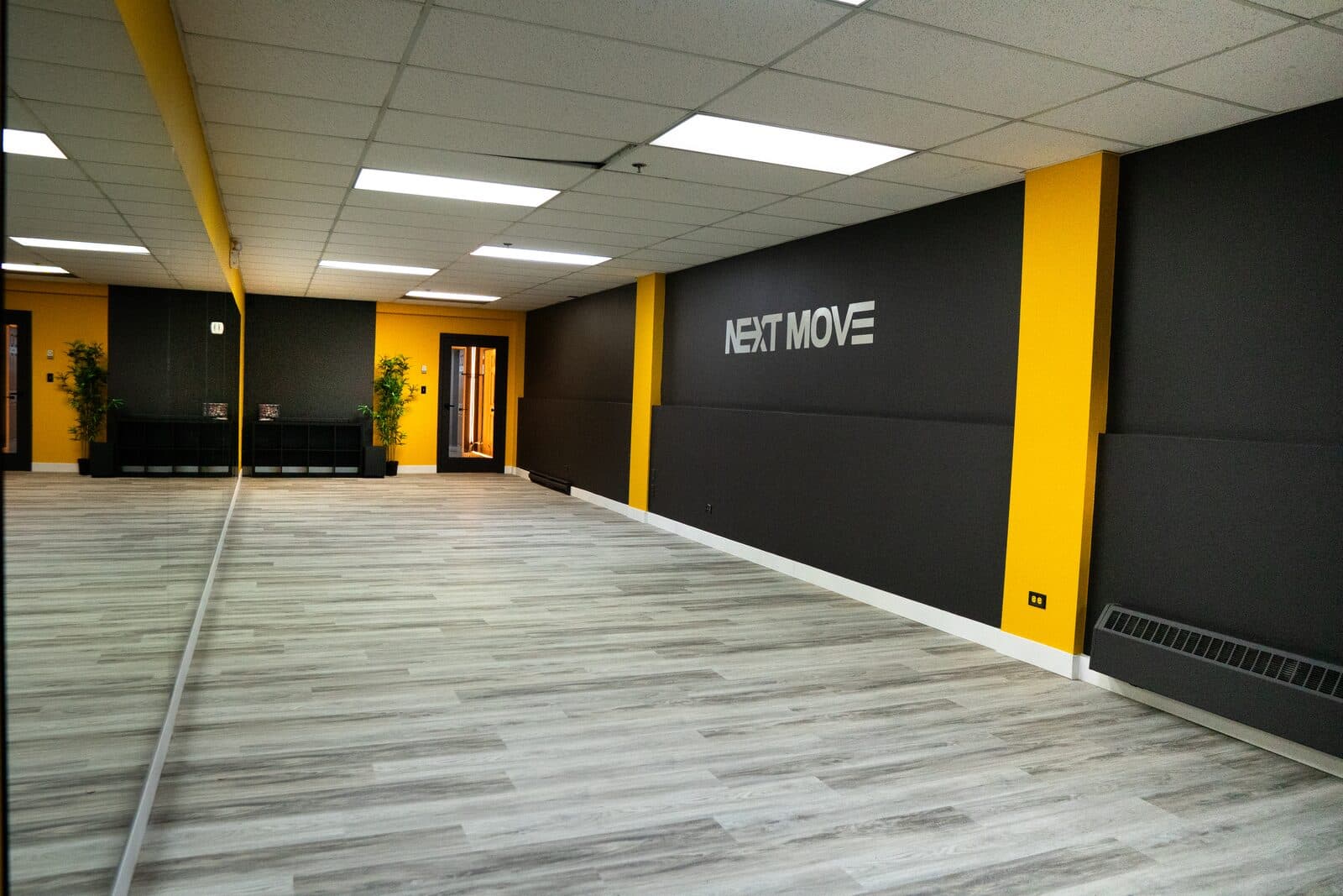 The Platform: an empty studio with a full-wall mirror, a grey wood floor and a black wall with the Next Move logo between two yellow pillars.