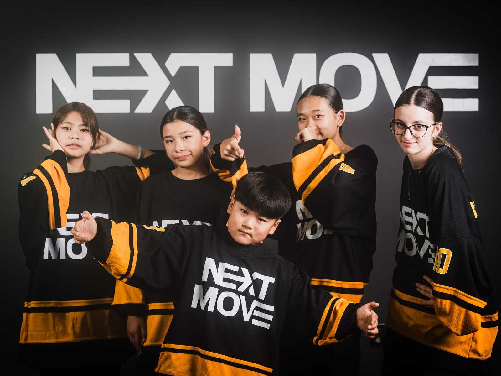 Five team dancers in black and yellow Next Move jerseys, posing together in front of the logo wall.
