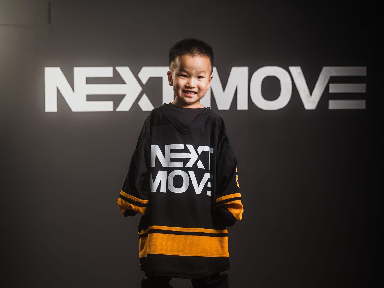 A young dancer in an oversized Next Move jersey, smiling in front of the studio's logo wall.