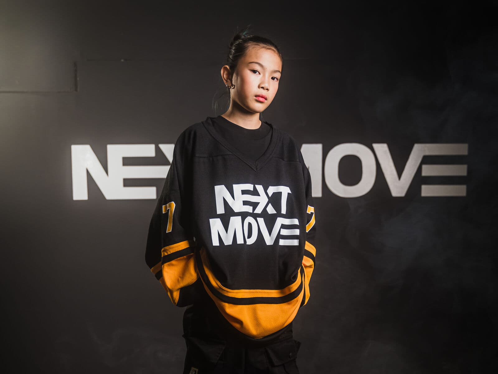 A young dancer in a Next Move jersey stands with hands in pockets, in front of the studio's logo wall.