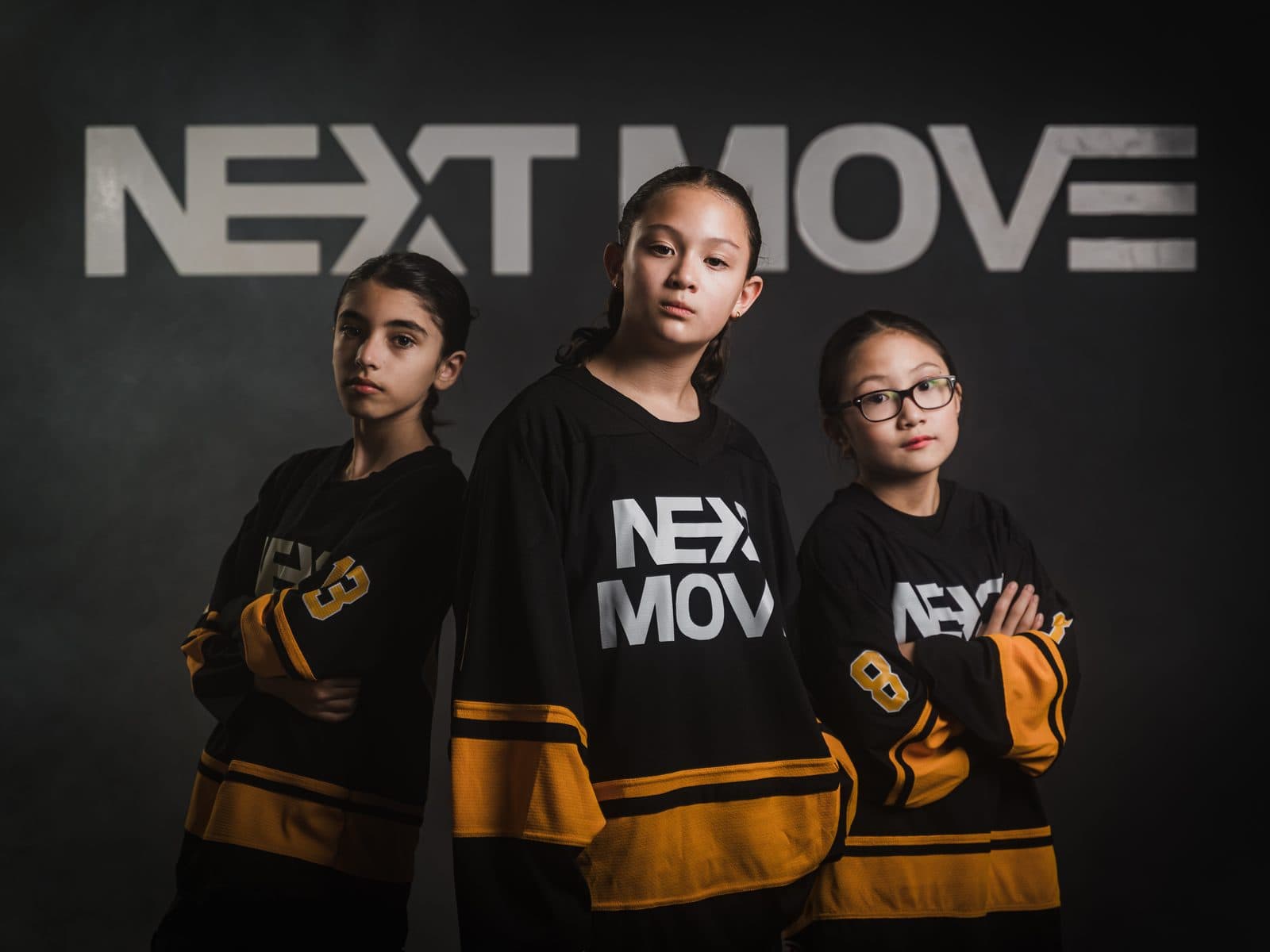 Three young dancers in Next Move jerseys, arms crossed, in front of the studio's logo wall.