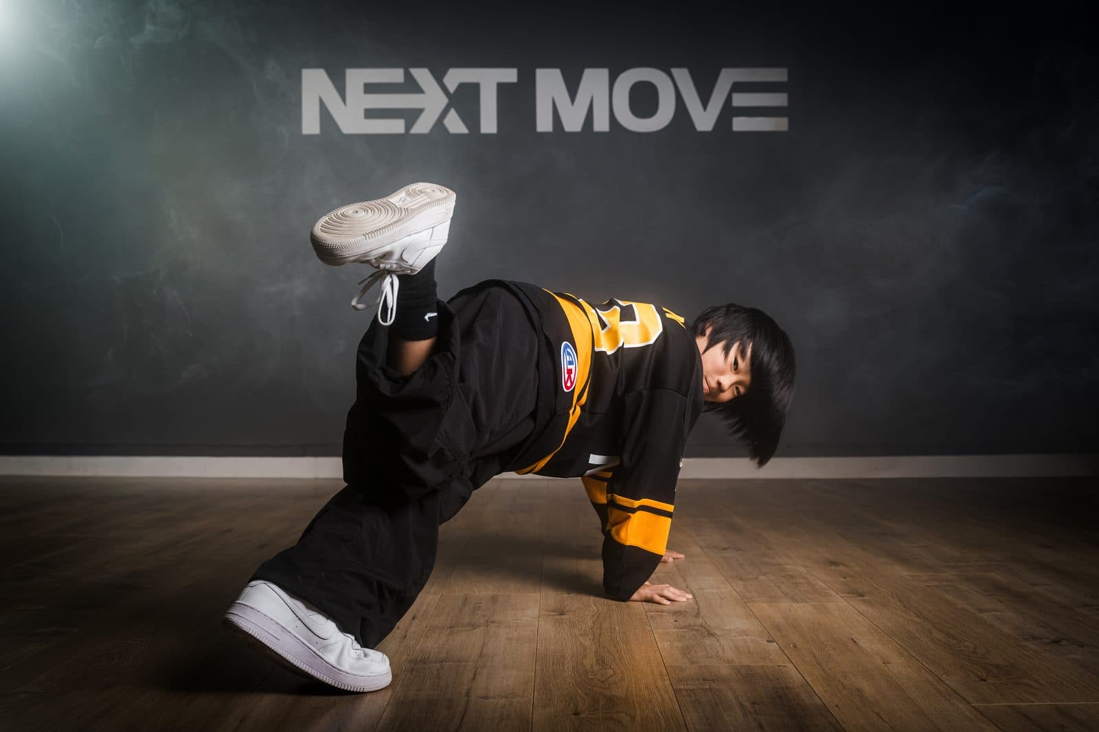 A young breaker holding a freeze on the studio floor, one foot in the air, under the Next Move logo.