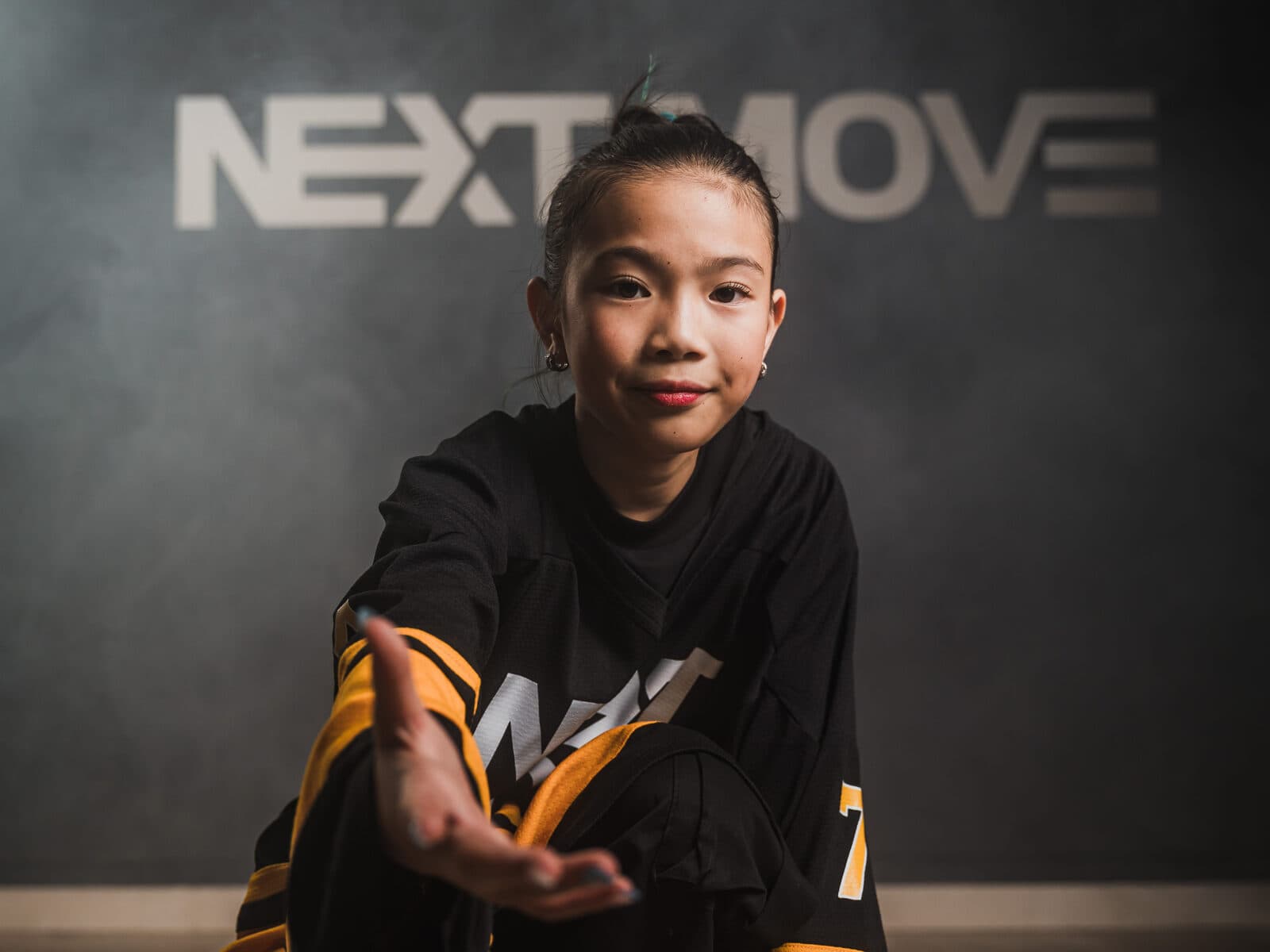 A young dancer in a Next Move jersey kneels in front of the studio's logo wall and reaches one hand toward the camera.
