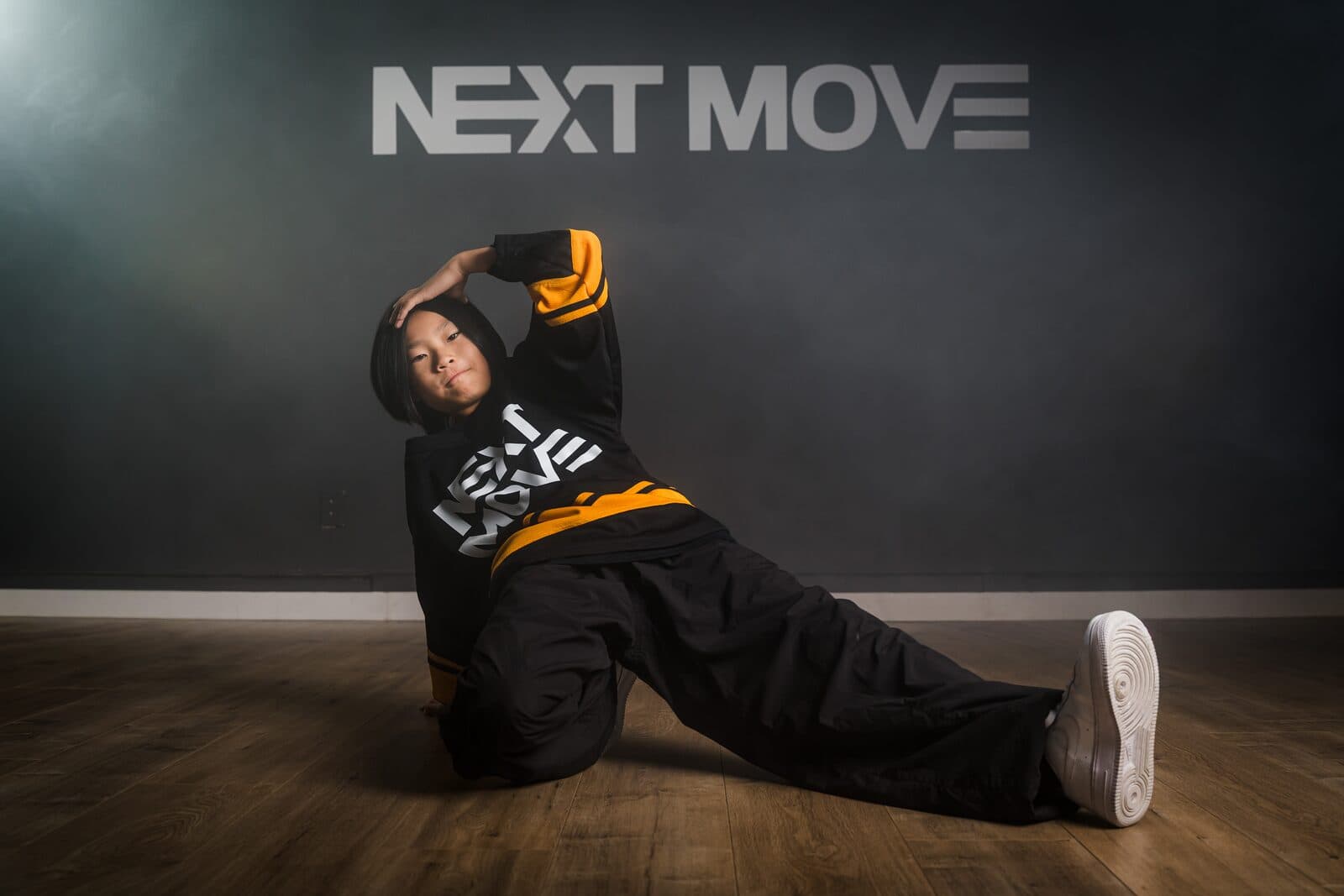 A young breaker kneels on one knee with one leg out and one hand on the head, under the Next Move logo.