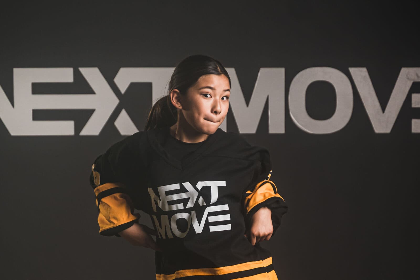 A young dancer in a Next Move jersey tilts the head with pressed lips and both hands at the hips, in front of the studio's logo wall.