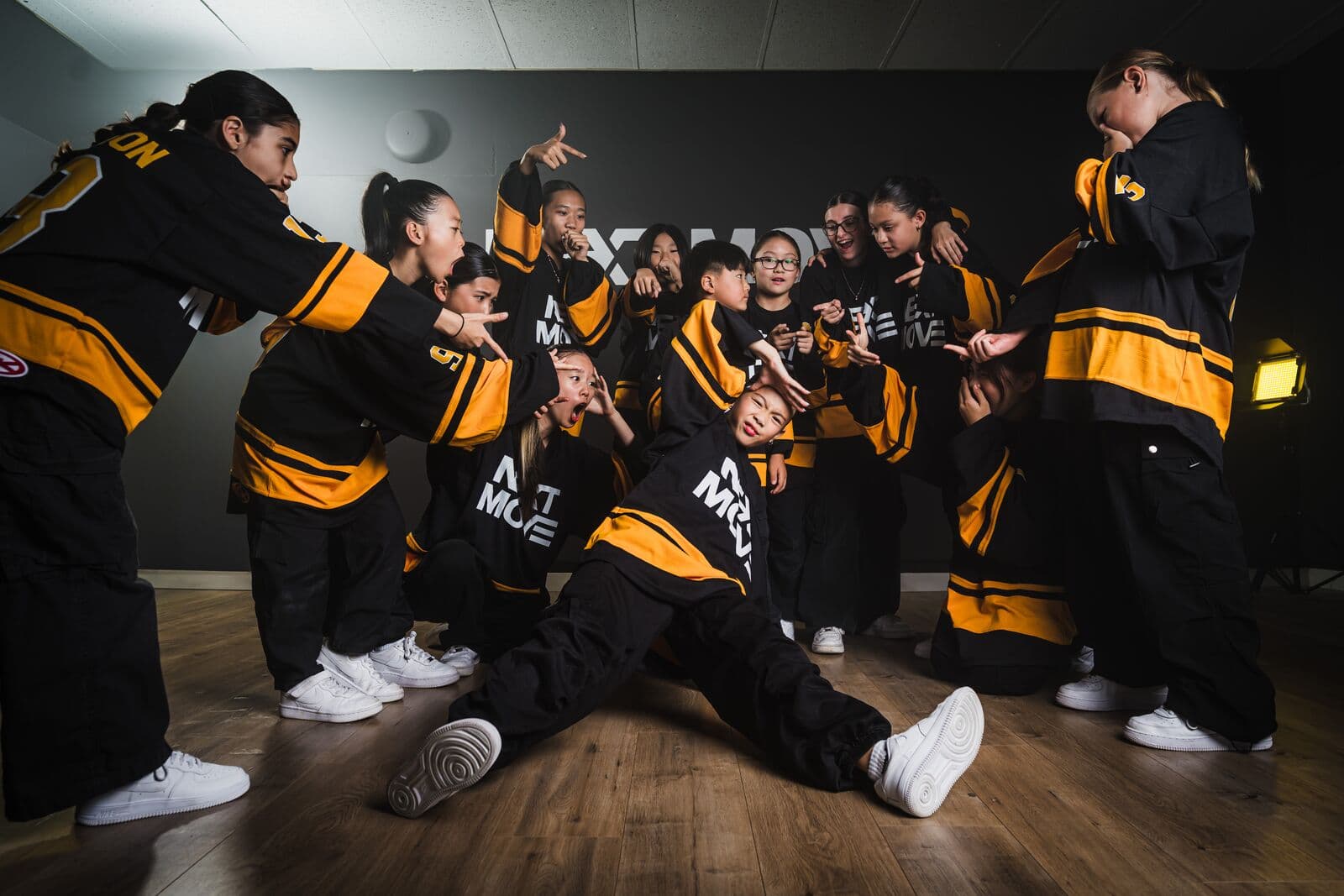 A young dancer sits in a wide split on the floor while a crew in Next Move jerseys points and reacts around them.