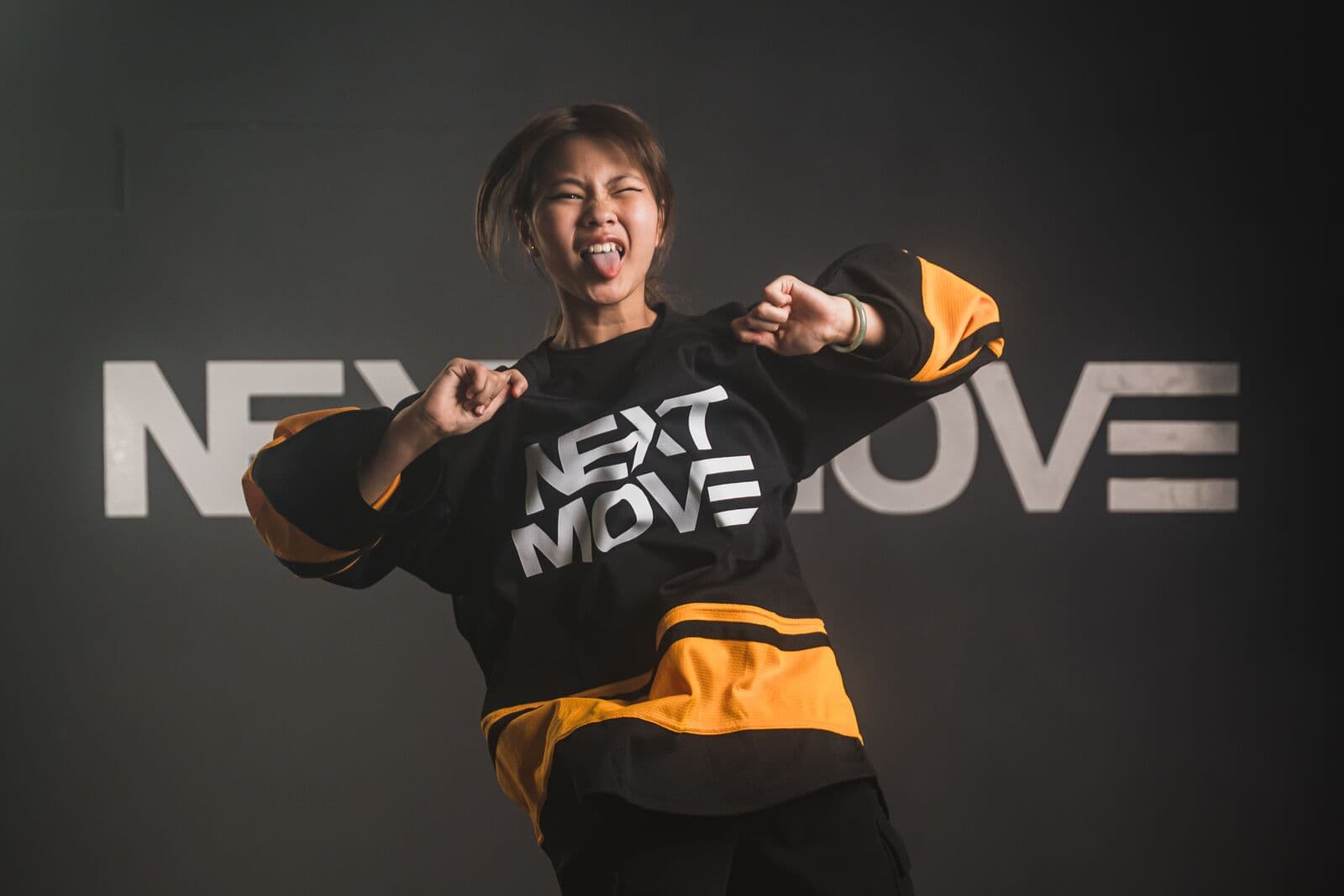 A young dancer in a Next Move jersey strikes a pose with both fists raised and sticks out the tongue.